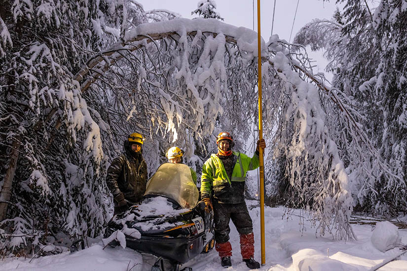 Three people stand next to a snowmobile in snow covered trees while one man holds a tall yellow pole