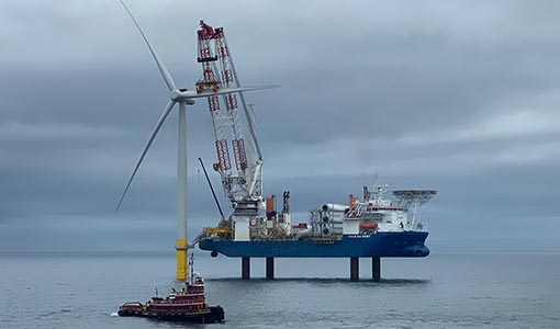 A crane on a large vessel installs a blade on an offshore wind turbine near a tugboat.
