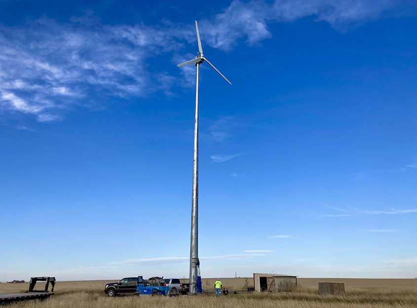 A wind turbine in a grassy field with people, vehicles, and equipment at its base