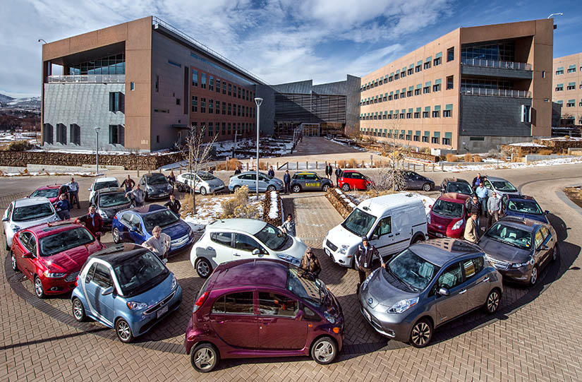 A group of people pose with their alternative fuel cars in front of a large office building.