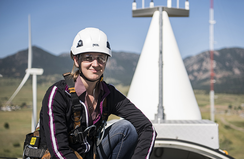 A person wearing a helmet and climbing gear sits atop a wind turbine.
