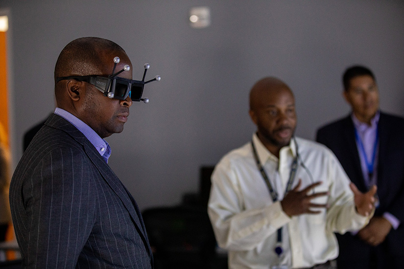 A man in the foreground wears VR goggles with two people in the background.