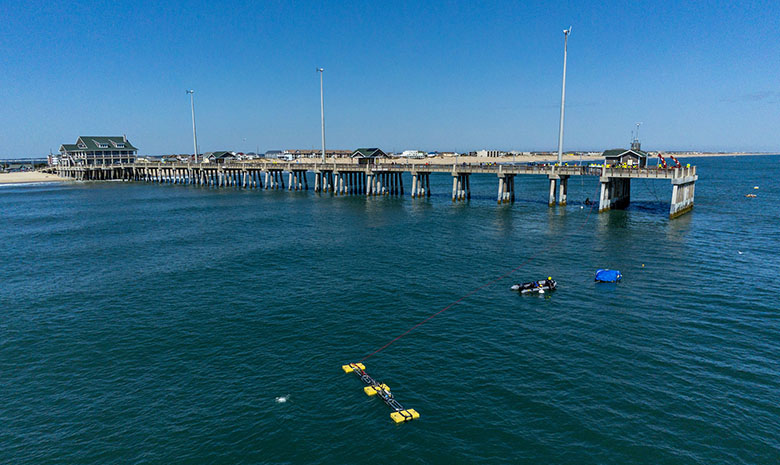 A diver moving away from a device that floats in the ocean next to a pier leading to a beach.