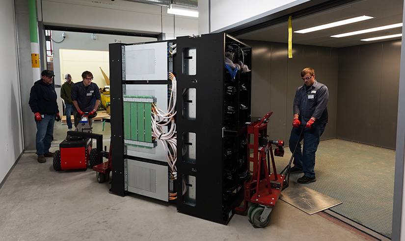 A group of people move a piece of the supercomputer via trolley cart into a room. 