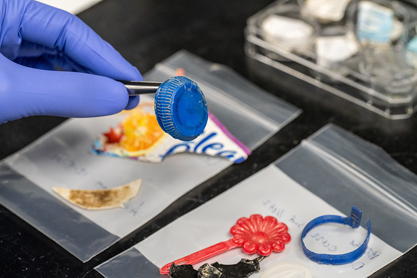A researcher uses tweezers to hold up a plastic bottle cap above a table with other bits of plastic.