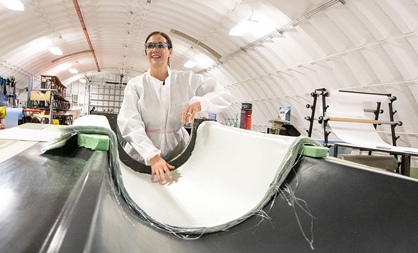 Robynne Murray wearing a jumpsuit and goggles and gesturing to an unfinished piece of a wind turbine