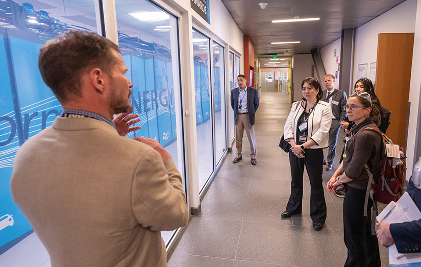 A group of seven people stand in a hallway, listening to one man.