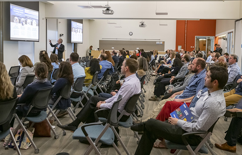 A crowded room of people watches a man with a PowerPoint presentation. 