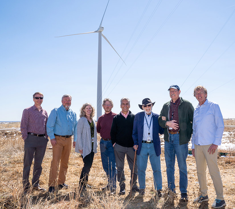 Eight people stand together outside under a wind turbine
