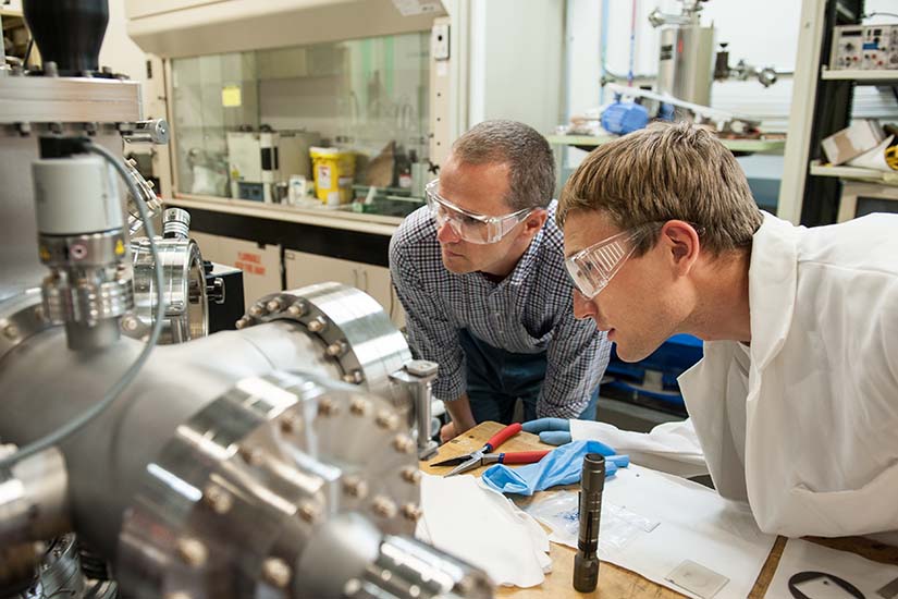 Two people wearing goggles lean over a lab bench to look at something inside a large metal device.