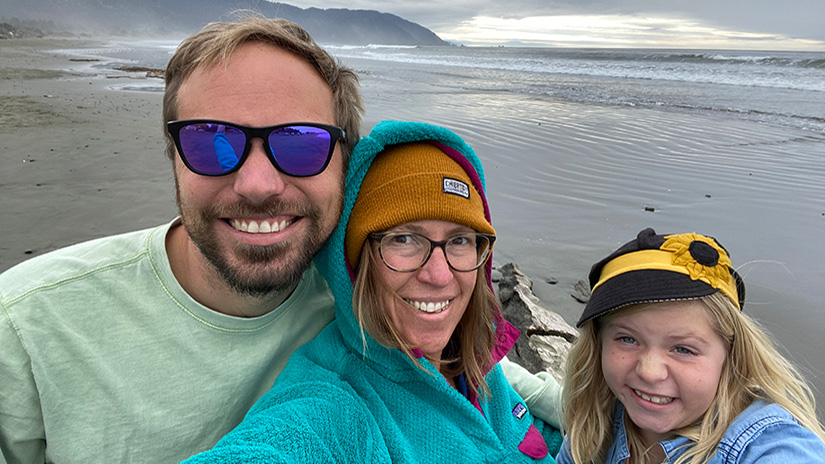 A man, woman, and young girl smiling in a selfie in front of the ocean with a hilly shoreline and gray skies.]