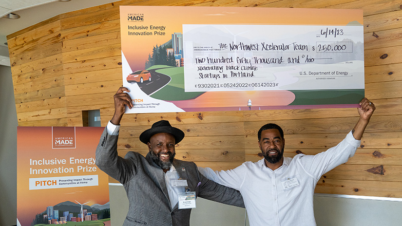 Two people hold up a giant check in front of a wood-paneled wall and a poster for the Inclusive Energy Innovation Prize PITCH event.