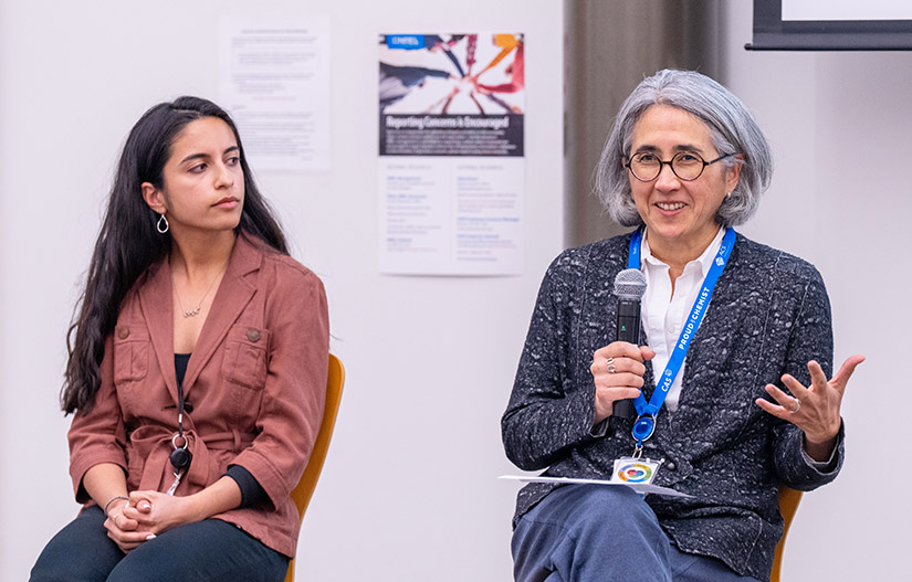 Two women sit on a panel, one speaking into a microphone.