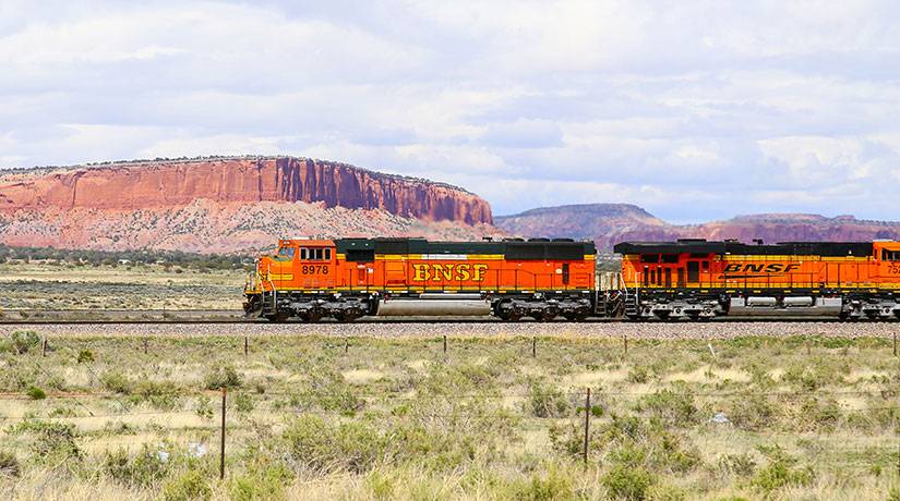An orange BNSF freight train crosses a flat green plain in front of a colorful desert mesa. 
