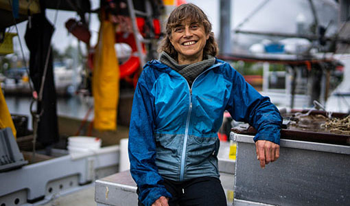 Linda Behnken, in a blue fishing jacket, smiles at the camera. Behind her is a colorful boatyard.