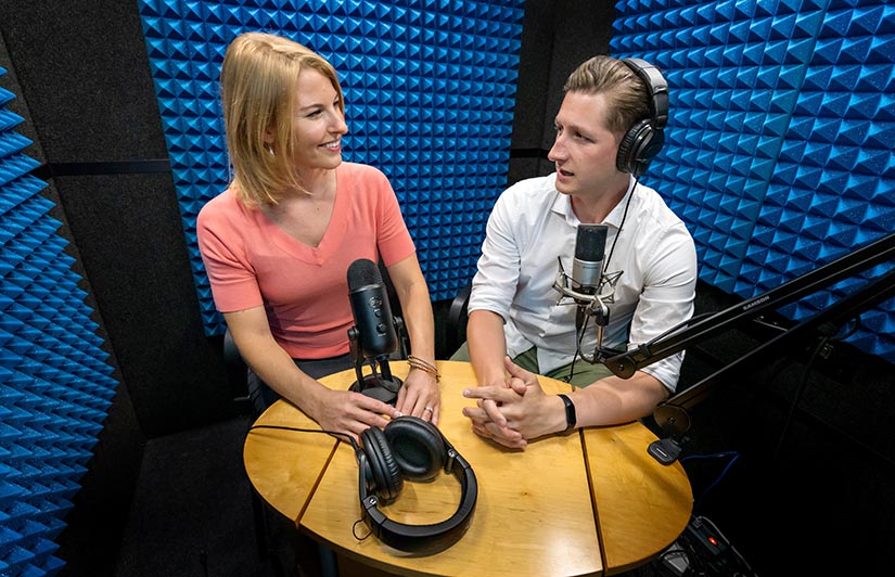 Two people sitting behind microphones in a soundbooth. 