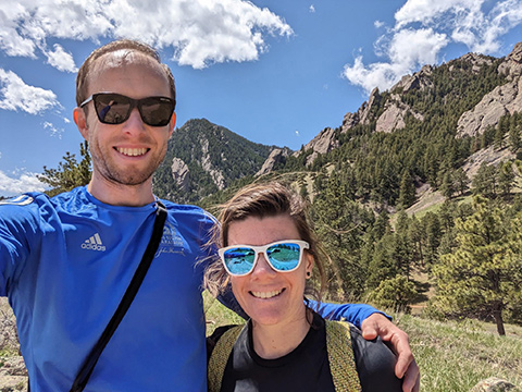 Two people smile for a selfie in front of a mountainous landscape.