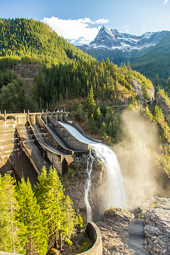 A large multi-level dam nestled in a steep tree-lined gorge, with a snowy mountain peak in the background.