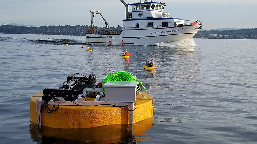 In the foreground is a round wave energy converter with metal piping and cables on top. Five attached buoys lead to a boat in the background.