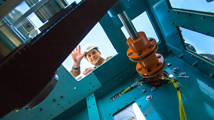 A man wearing a hard hat waves through an opening at the top of a metal structure containing a turbine.