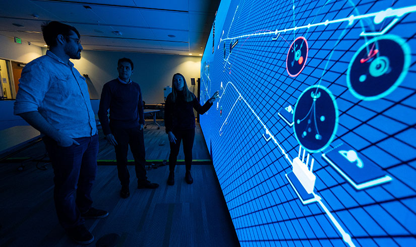Three people stand in front of a large computer screen filled with small blue squares and other data.