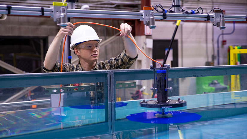 A man in a hard hat moves a cable attached to a small device floating in a large tank of water.