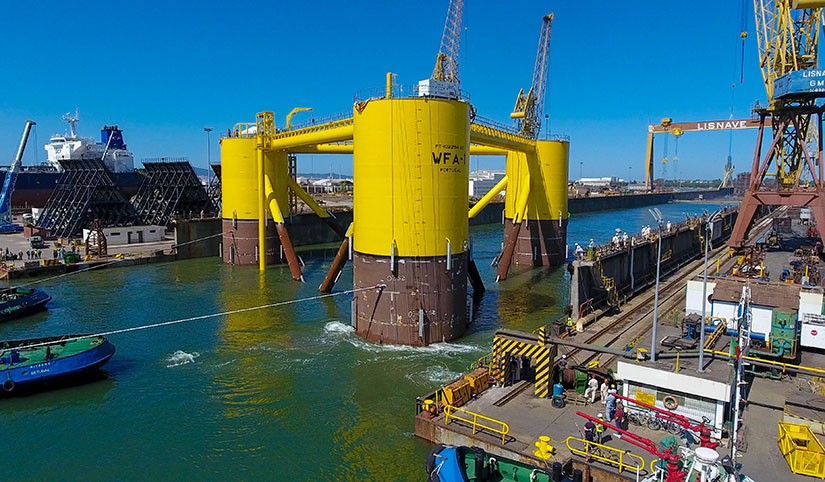 Three connected columns being towed by a boat from a water channel next to harborside cranes