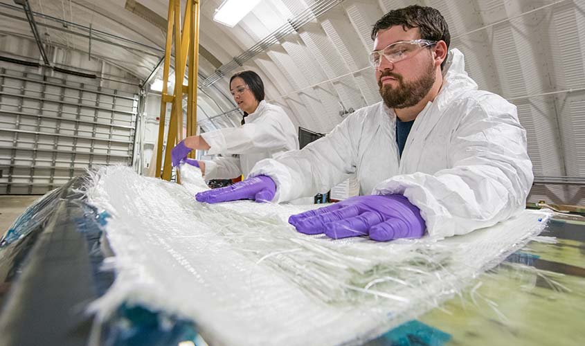 Two people wearing safety goggles, lab coats, and gloves placing composite materials in a turbine blade mold. 