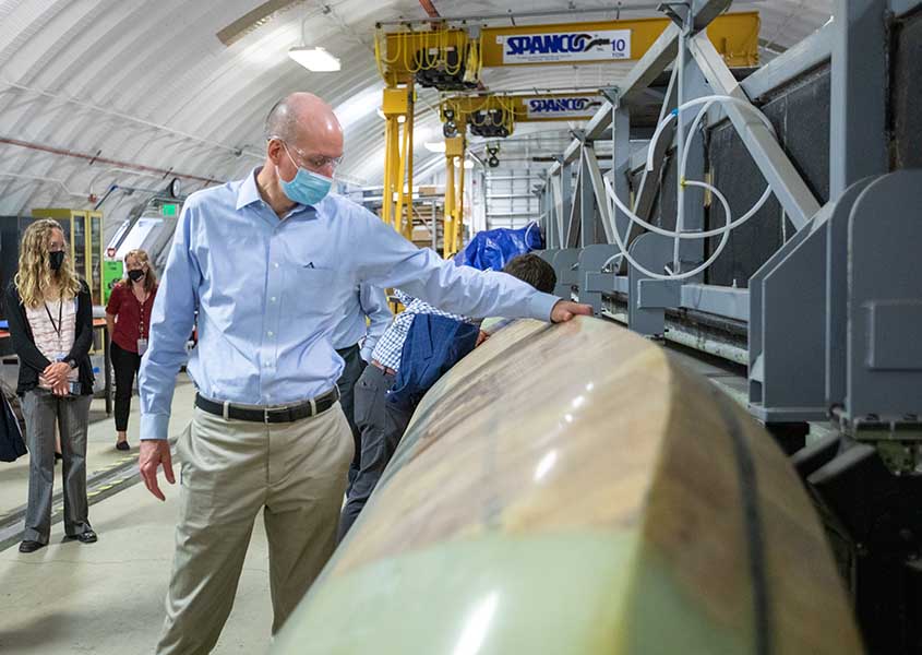 A man in a mask touching a long turbine blade. Several people can be seen behind him in the research facility.