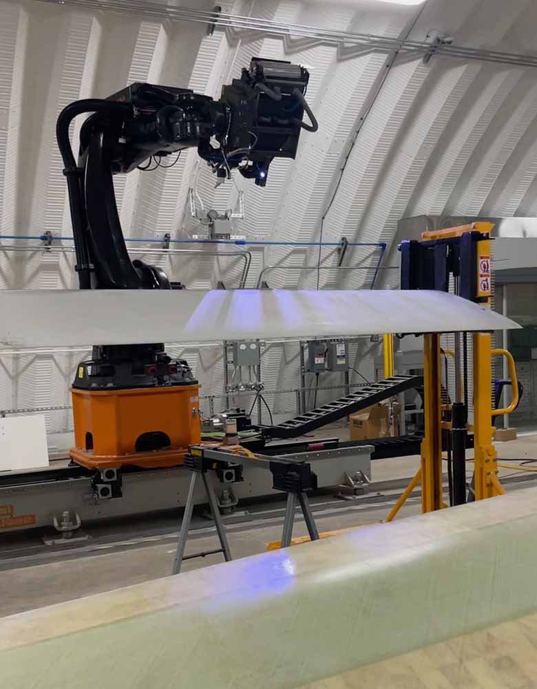 A large robotic arm shooting lasers on a wind turbine blade propped on stilts in a facility