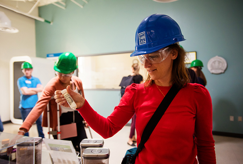 A woman in a red shirt and wearing a hardhat holds up a vial with a dollar bill and salt in it.