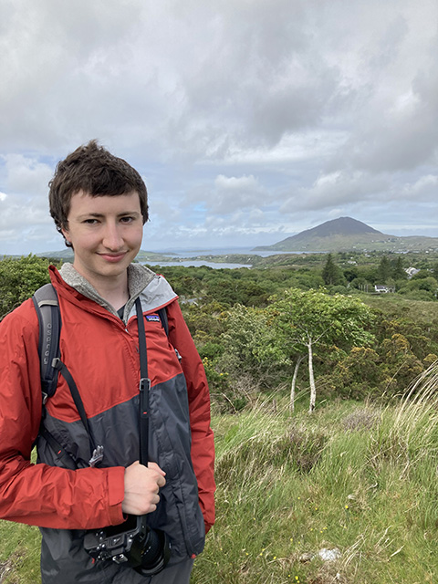 A person in a rain jacket poses while hiking in front of a mountain.