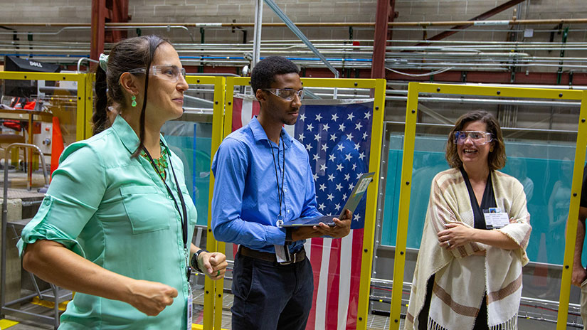 Three people in safety glasses talk in front of a wave tank draped with the U.S. flag.