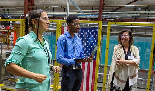 Three people in safety glasses talk in front of a wave tank draped with the U.S. flag.