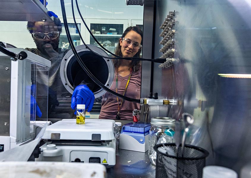 : Two people in protective goggles use a glove box to put a vial on a scale in a lab