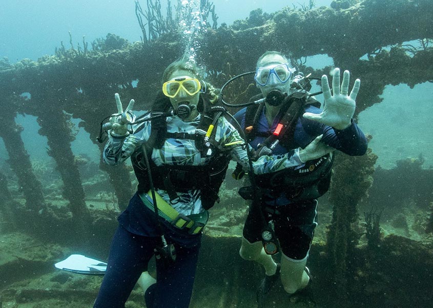 Two people under water wearing scuba gear in front of wreckage.