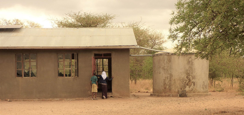 Two children walking into a small building with pipes extending from the roof to a water tank nearby.