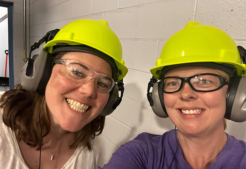 Two people wearing construction hats and ear protectors smile. 