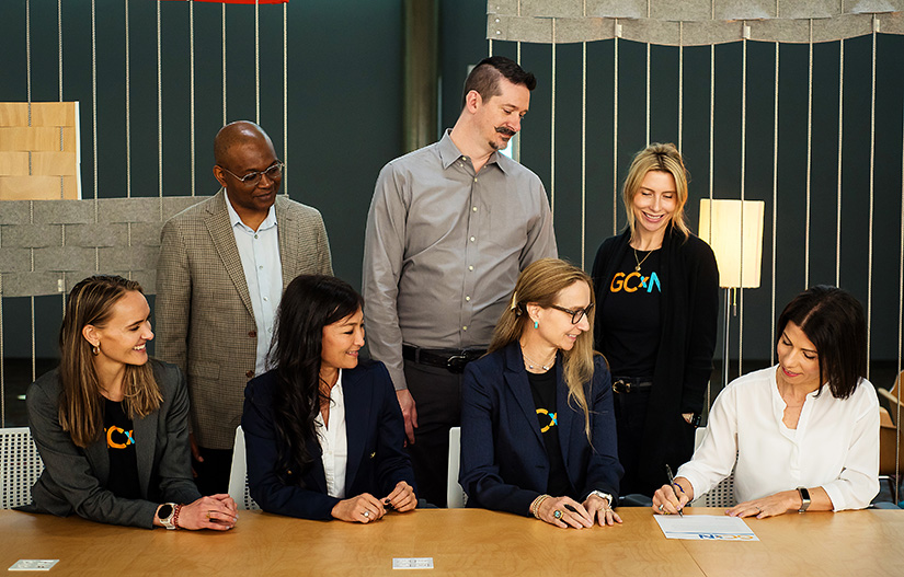 A few people sit at a table, with a few others standing behind them, watching a woman at the end of the table signing a piece of paper. Several of the people are wearing GCxN shirts.