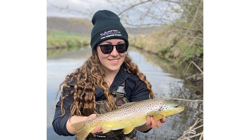 Natalie Coash wearing waders and holding a fish while standing nex to a river.