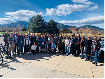 A group of people stand outside, with spruce trees, buildings, mountains, and partly cloudy skies behind them. 