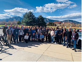 A group of people stand outside, with spruce trees, buildings, mountains, and partly cloudy skies behind them.
