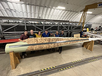 Seven people stand behind a wind turbine blade propped up on stands inside a hangar.