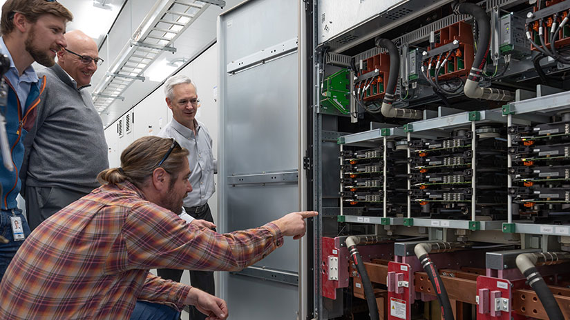 Four people look at electronic switches inside a machine.