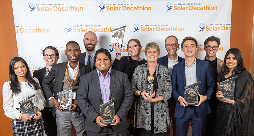 Eleven people stand in front of a photo backdrop with the Solar Decathlon logo holding seven trophies.