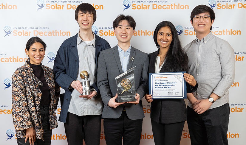 Five people stand in front of a photo backdrop with the Solar Decathlon logo holding two trophies and a certificate.