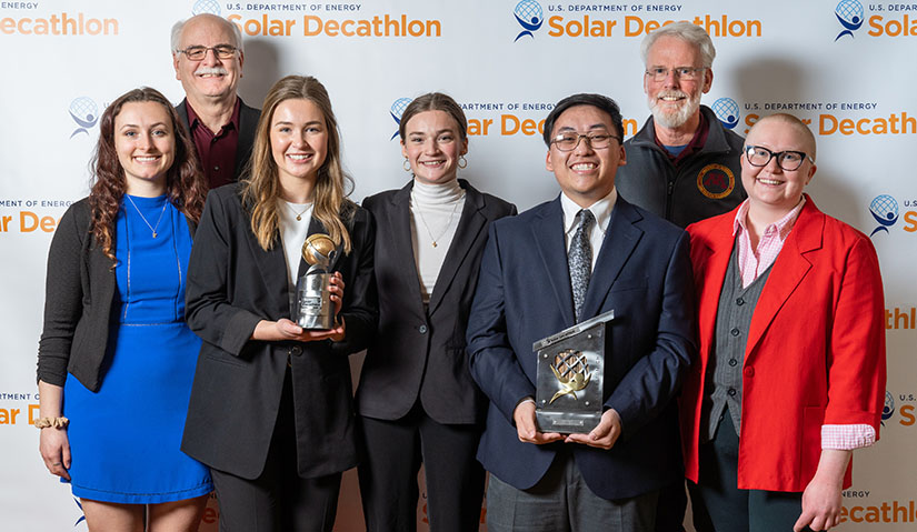 Seven people pose in front of a photo backdrop with the Solar Decathlon logo holding two trophies.