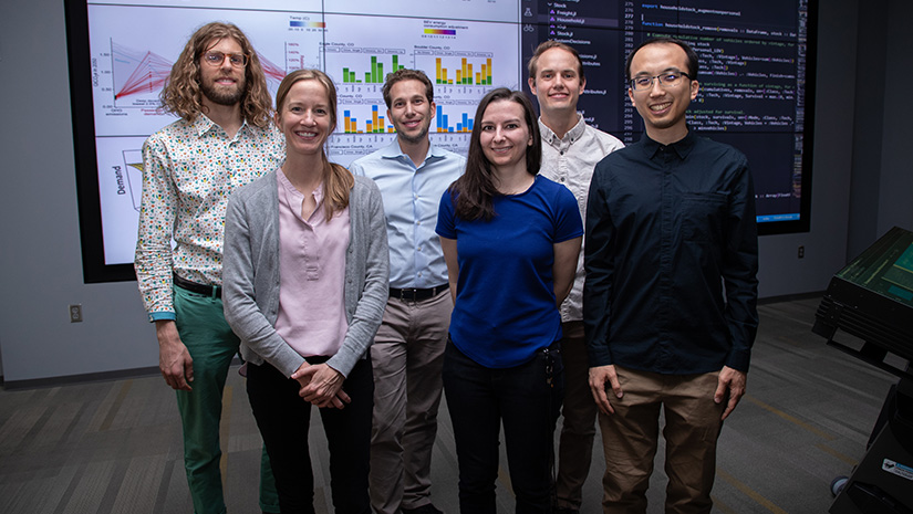 A group of six people standing in front of a large-screen visual display showing maps and charts.