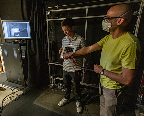 Two researchers stand with a hand-held solar panels connected to testing equipment while examining results on a screen.