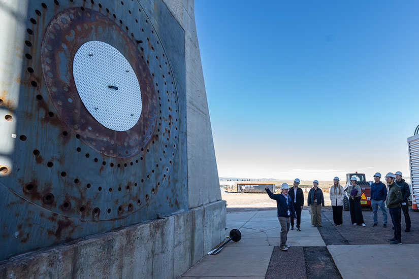 A group of people stand outside in front of a large structure, listening to their tour guide.